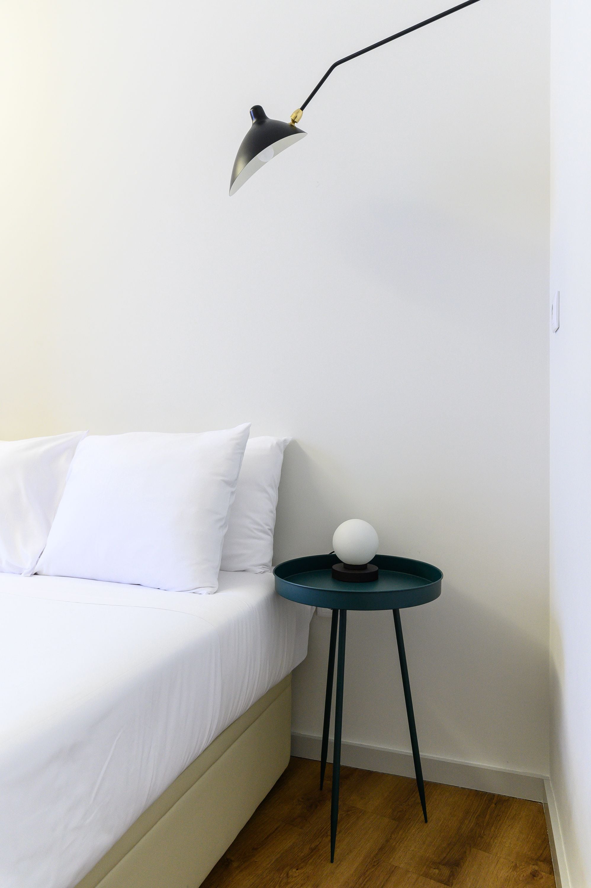 A bedroom with a bed, white bedding, a small round table with a white spherical object, and a modern floor lamp.