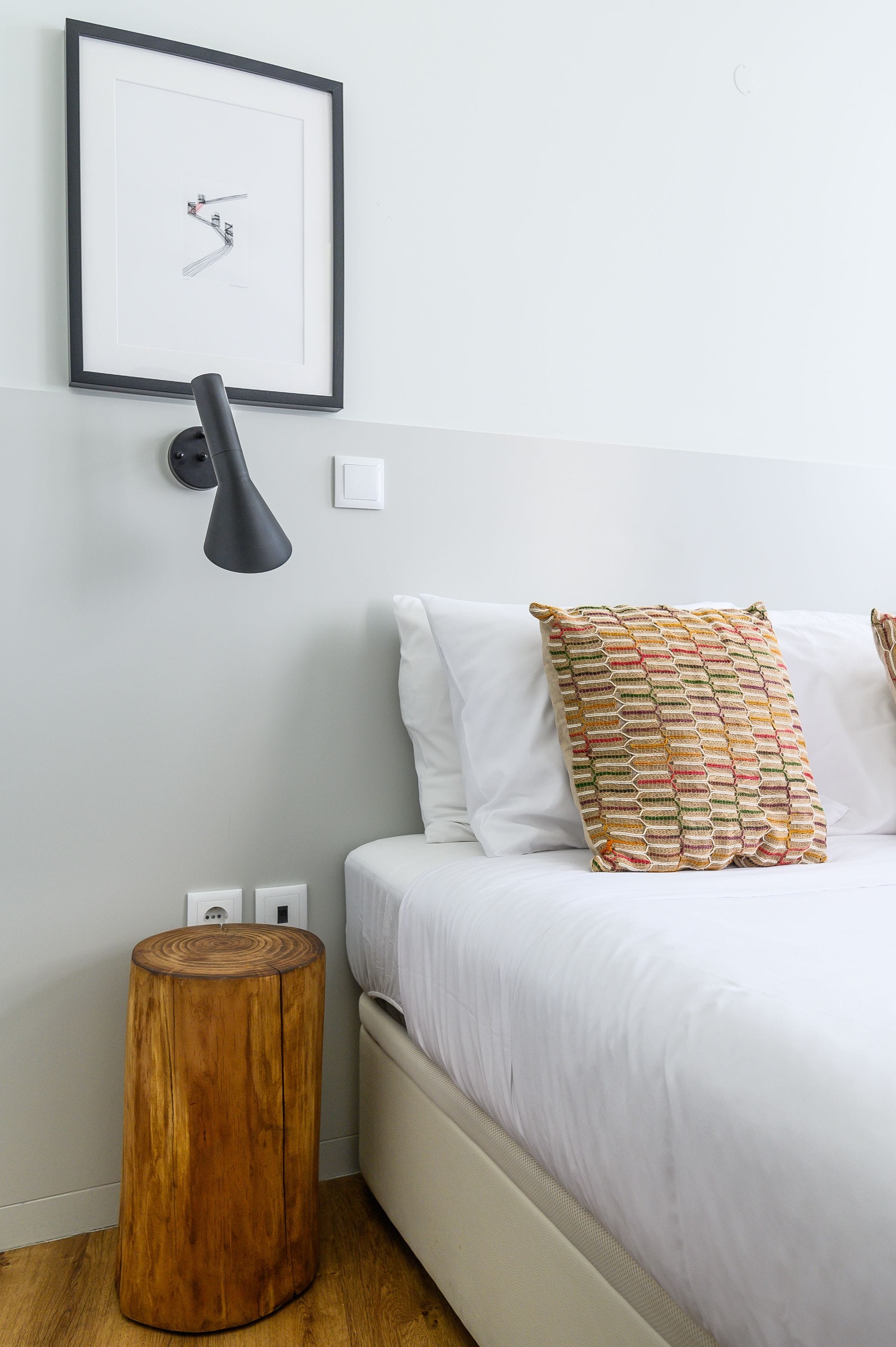 A bedroom with a bed, a bedside lamp, a framed picture on the wall, and a wooden stool.