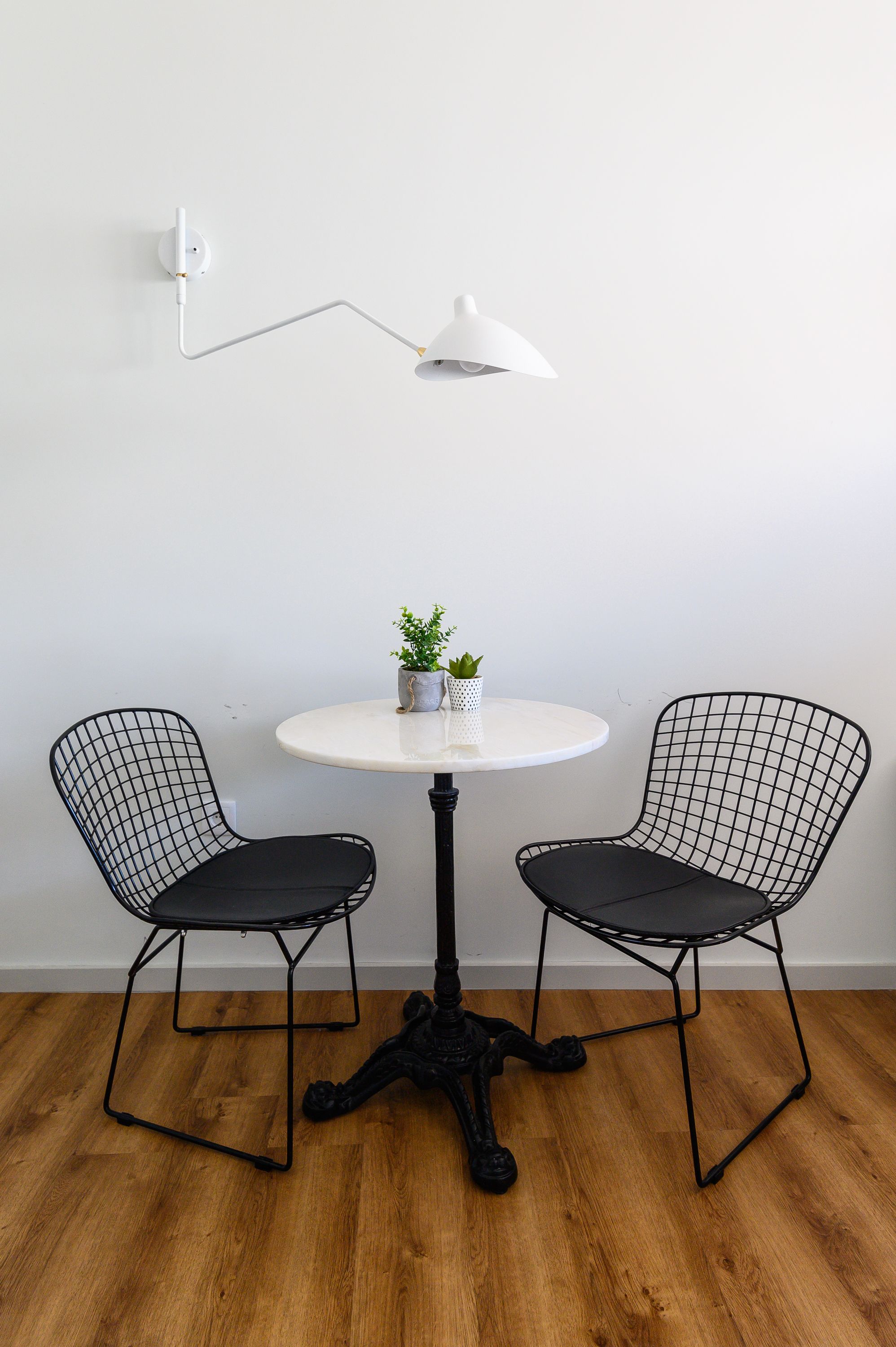 A small round table with two black wireframe chairs, a white floor lamp, and a potted plant on the table.