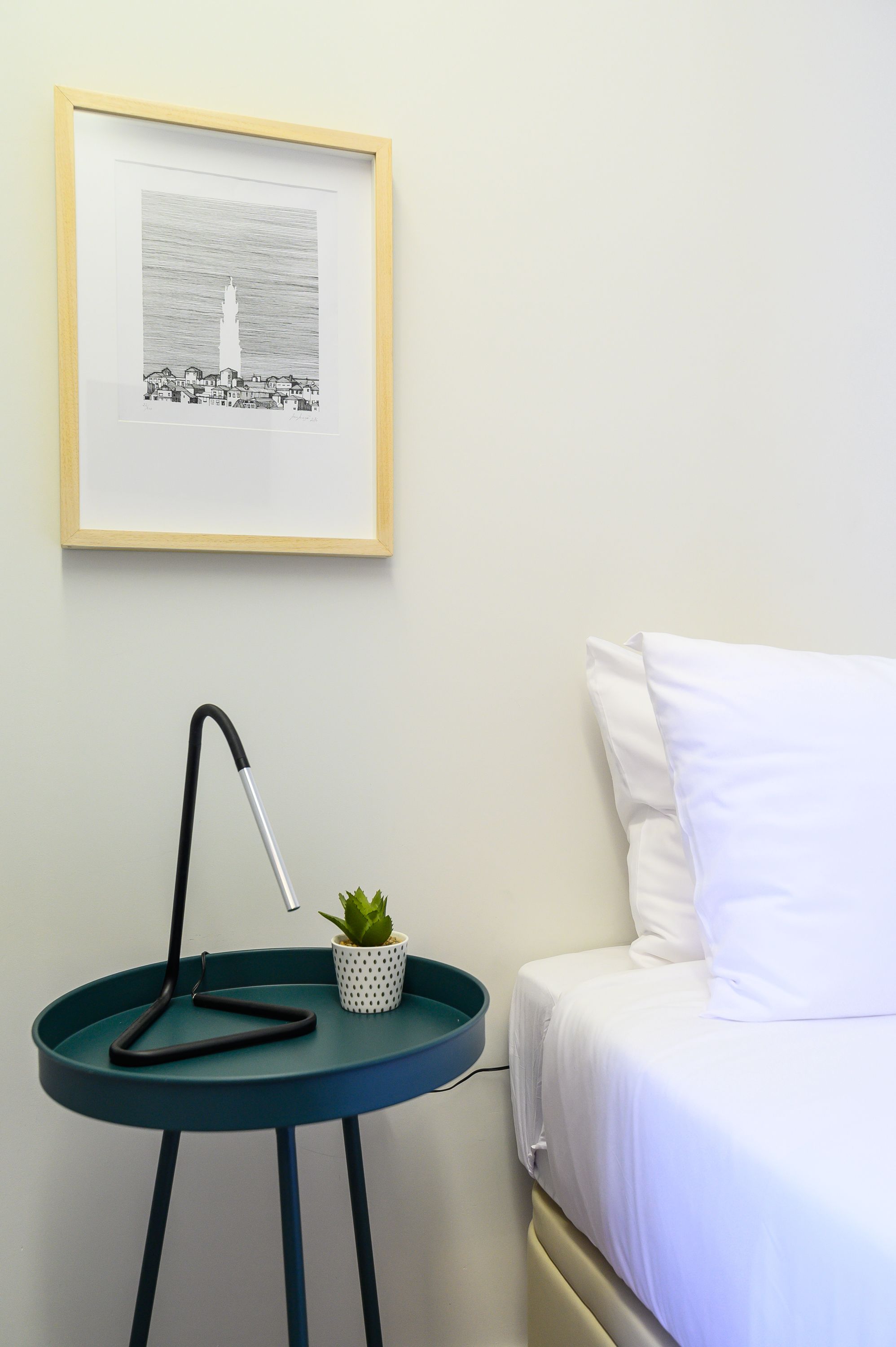 Quarto bem arrumado com uma cama, uma mesa de cabeceira com abajur, um quadro na parede e um pequeno vaso com planta.