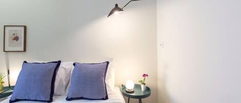 A neatly made bed with two pillows, a bedside table with a lamp, a small potted plant, and a framed picture on the wall.