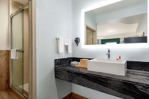 Un baño con cabina de ducha de vidrio, un lavamanos blanco sobre encimera de mármol negro y un espejo sobre el lavamanos.