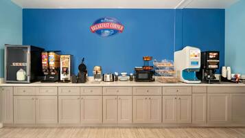 Una barra de desayuno con una pared azul, un letrero de Baymont Breakfast Corner, un refrigerador, cajas de cereales, un microondas, una cafetera y diversos productos para el desayuno.