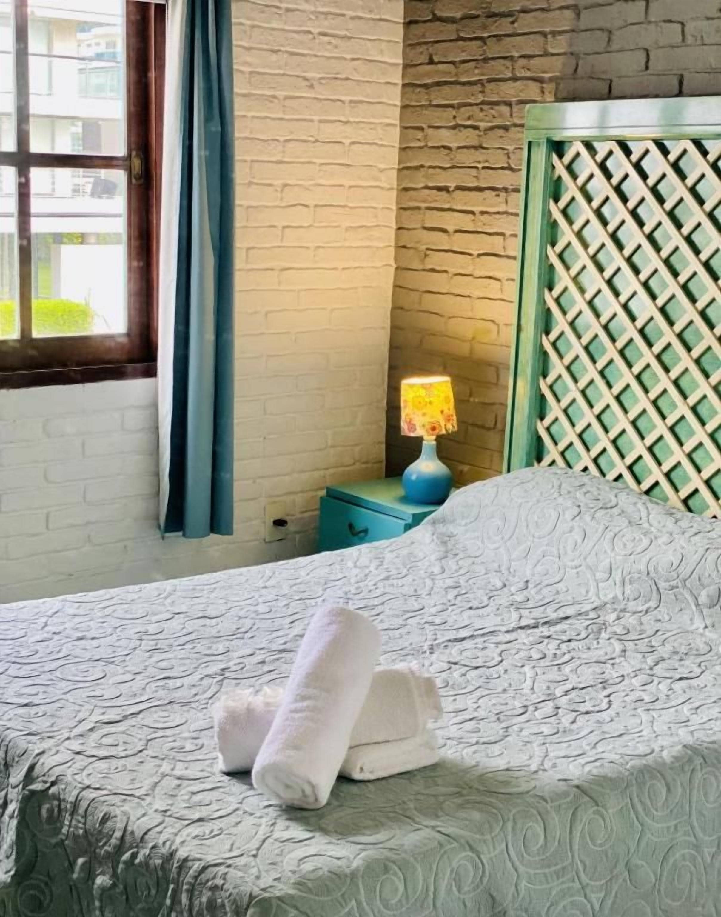 Quarto de hotel com cama, mesa de cabeceira com abajur e janela com cortinas.