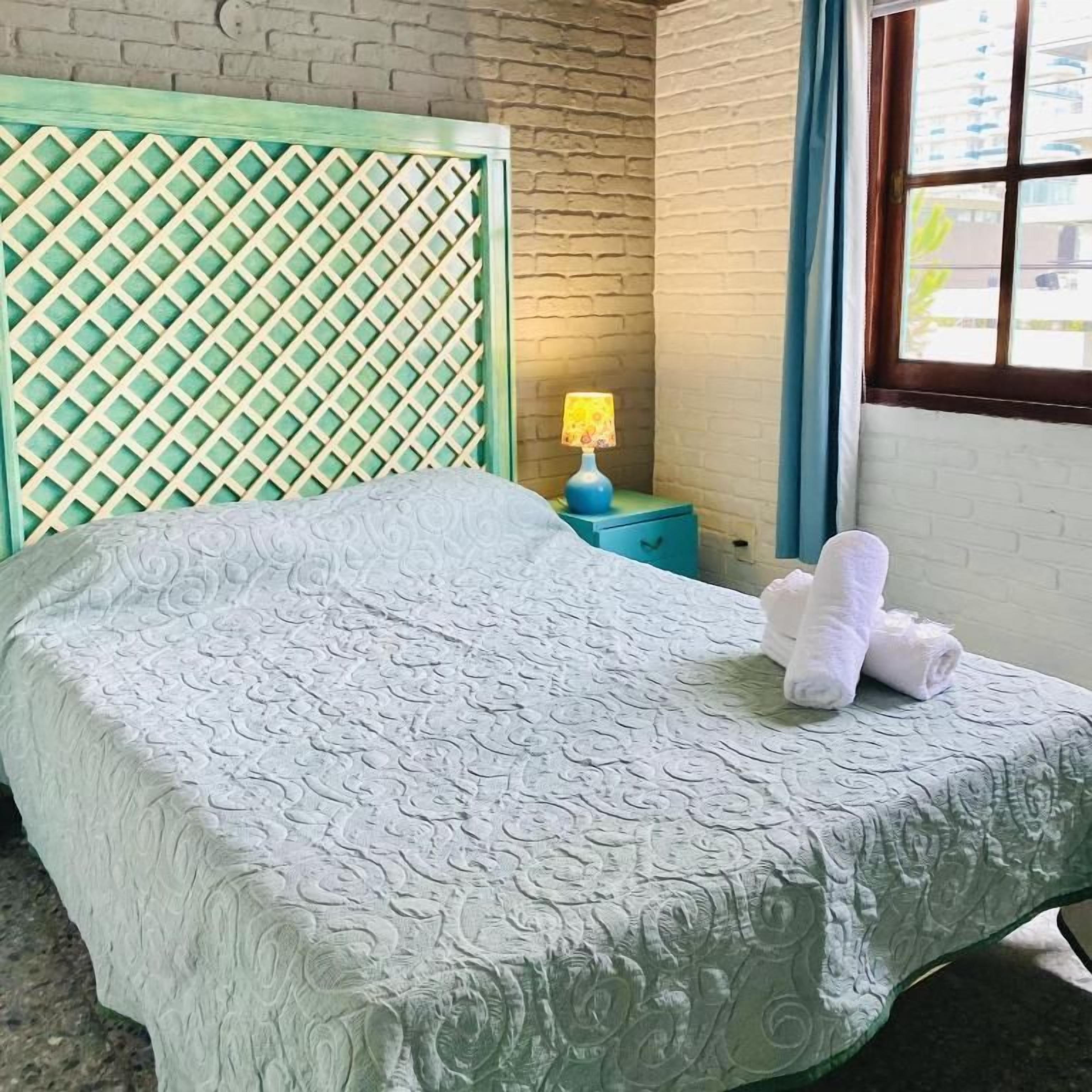 A bed with a patterned bedspread, a headboard with a lattice design, a nightstand with a lamp, and a window with curtains.