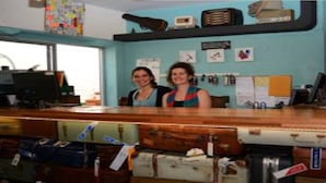A reception desk with two individuals standing behind it, a wall with framed pictures, and a large orange figure on the right. - El Viajero Hostels & Suites - Punta del Este (Punta del Este)