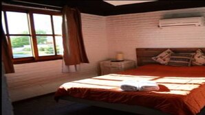 A bedroom with a wooden bed, a window with curtains, a bedside table with a lamp, and a wall-mounted air conditioner. - El Viajero Hostels & Suites - Punta del Este (Punta del Este)