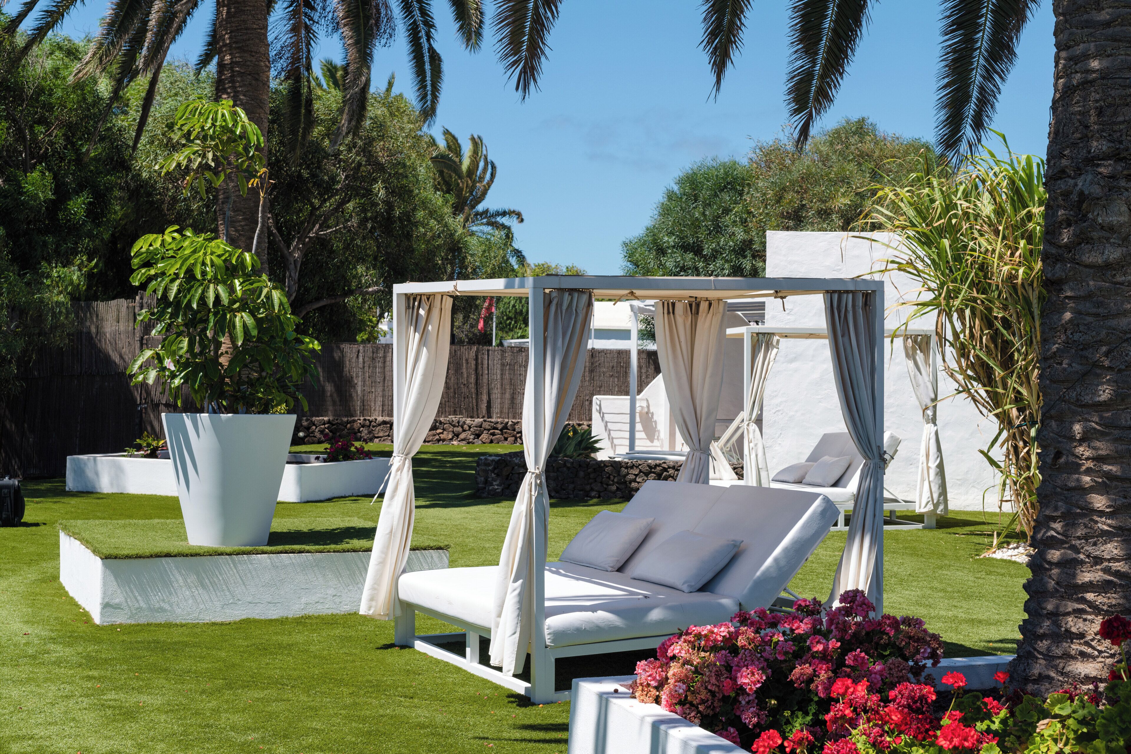 Een tuin met een wit hemelbed, een witte lounge stoel en een grote plantenbak vol bloemen.