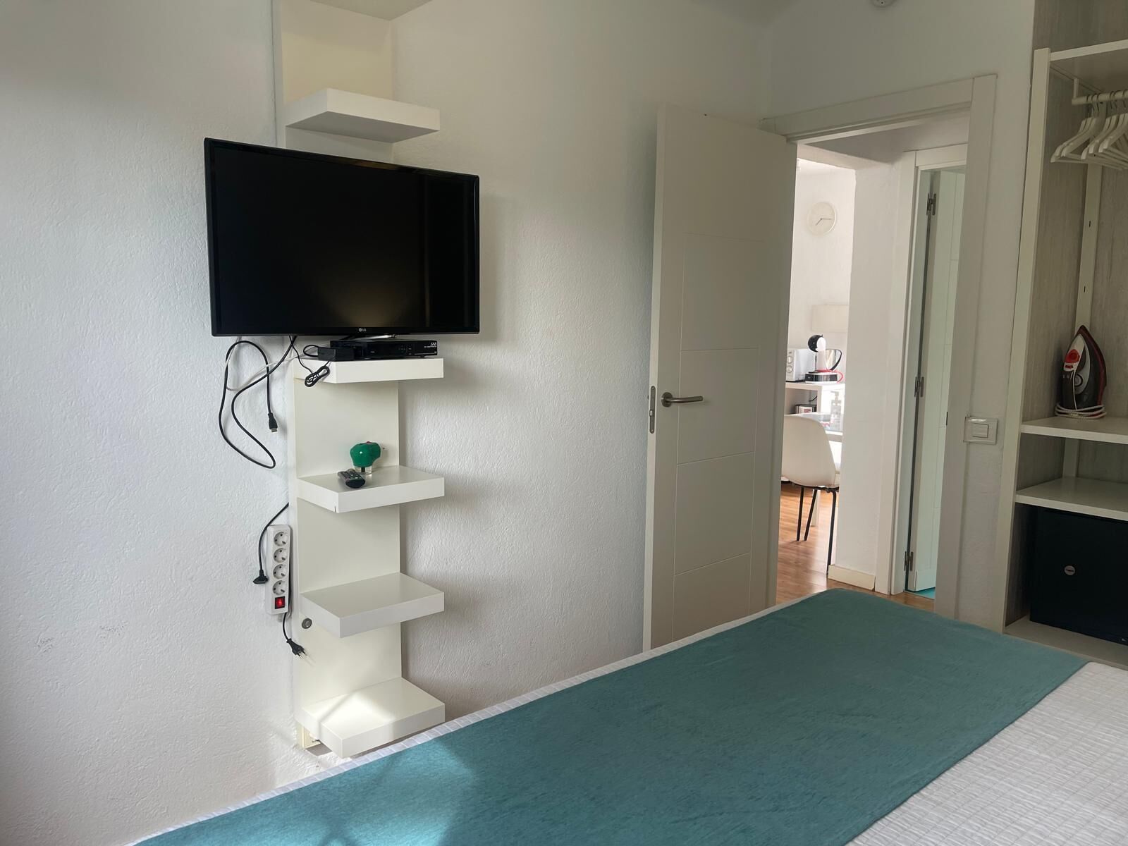 Un dormitorio con una cama, un televisor montado en una pared blanca y una puerta que da a otra habitación.