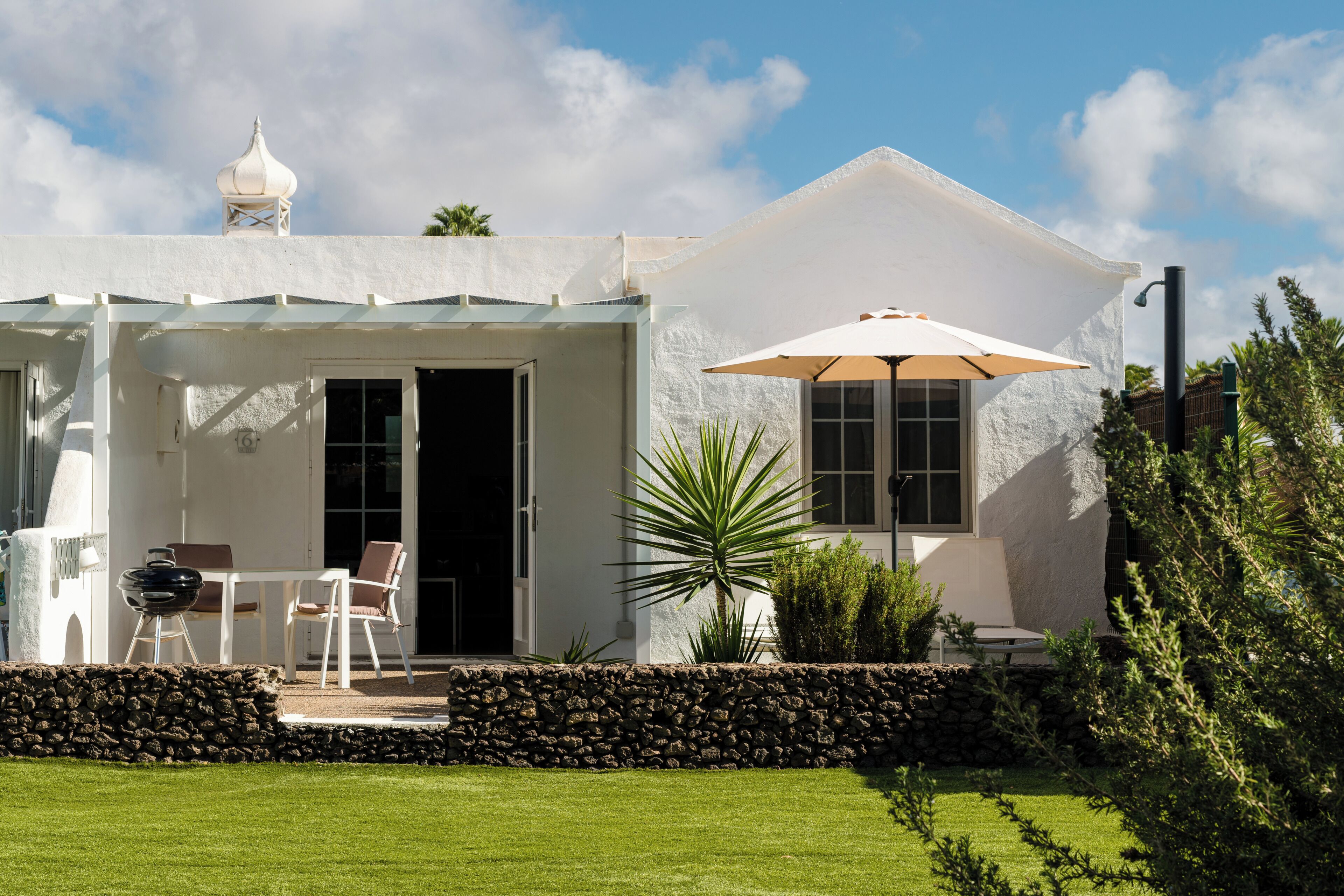 A white building with a covered patio, a stone wall, and a lawn.
