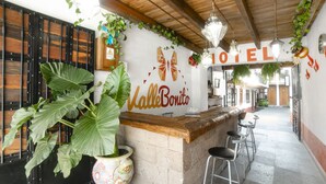 Zona de recepción de un hotel con mostrador de madera, una planta grande en maceta y taburetes de bar.