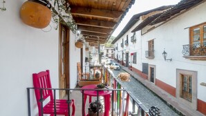 Un balcón con sillas rojas y una mesa, con vistas a una calle estrecha flanqueada por edificios.