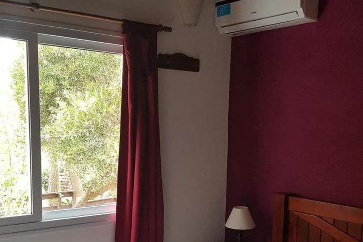 A bedroom with a bed, a window with curtains, a nightstand with a lamp, and an air conditioning unit on the wall.
