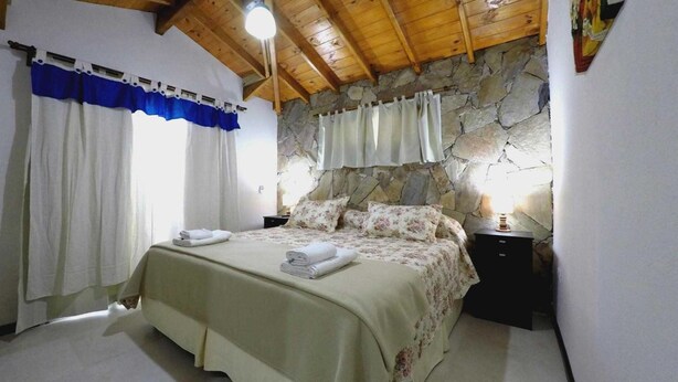 A bedroom with a stone wall, a bed with floral bedding, and two bedside tables with lamps. - Cabañas Victoria (Mar de Las Pampas)