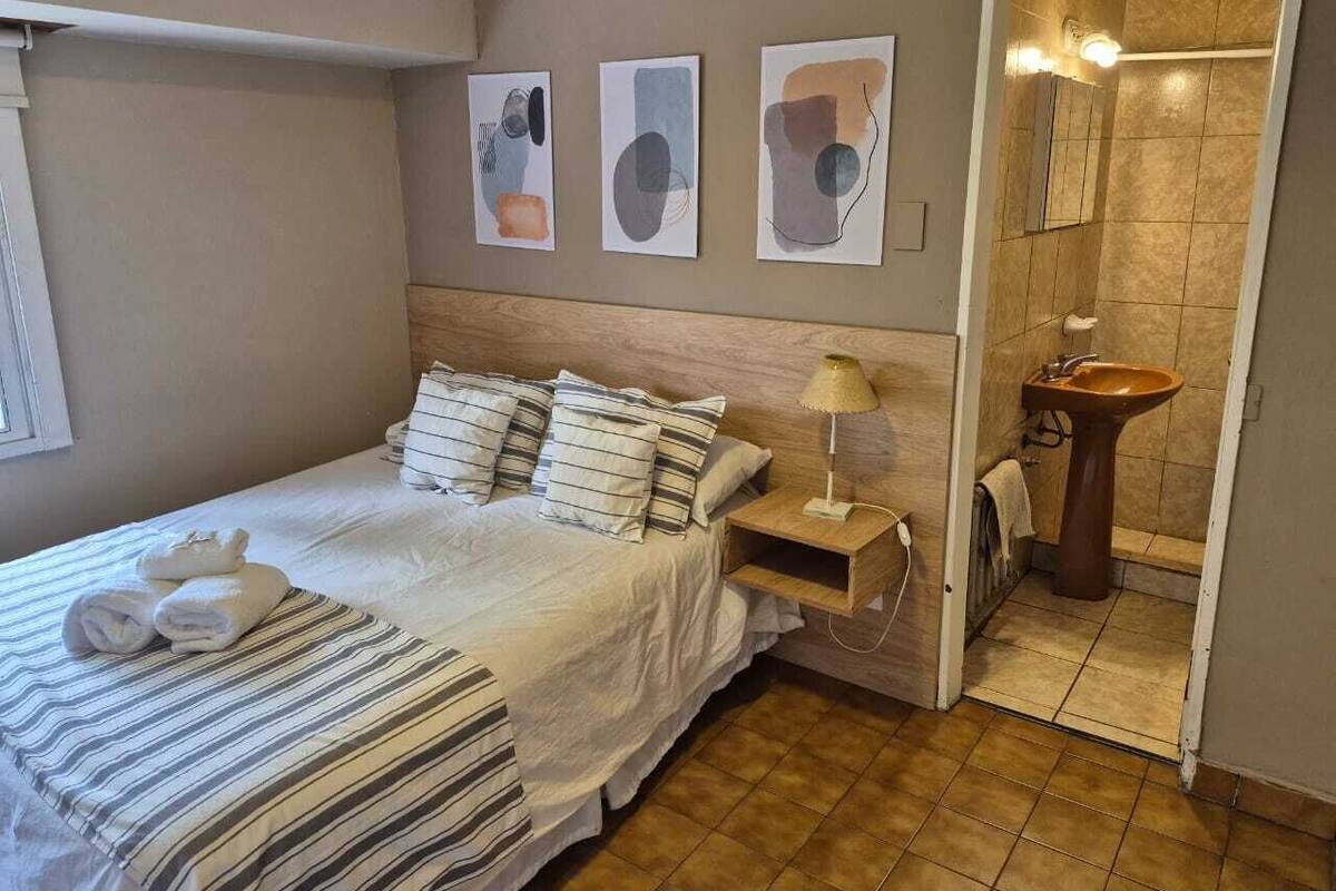 Een slaapkamer met een bed, kussens, een nachtkastje met een lamp, en een badkamer met een wastafel en spiegel.