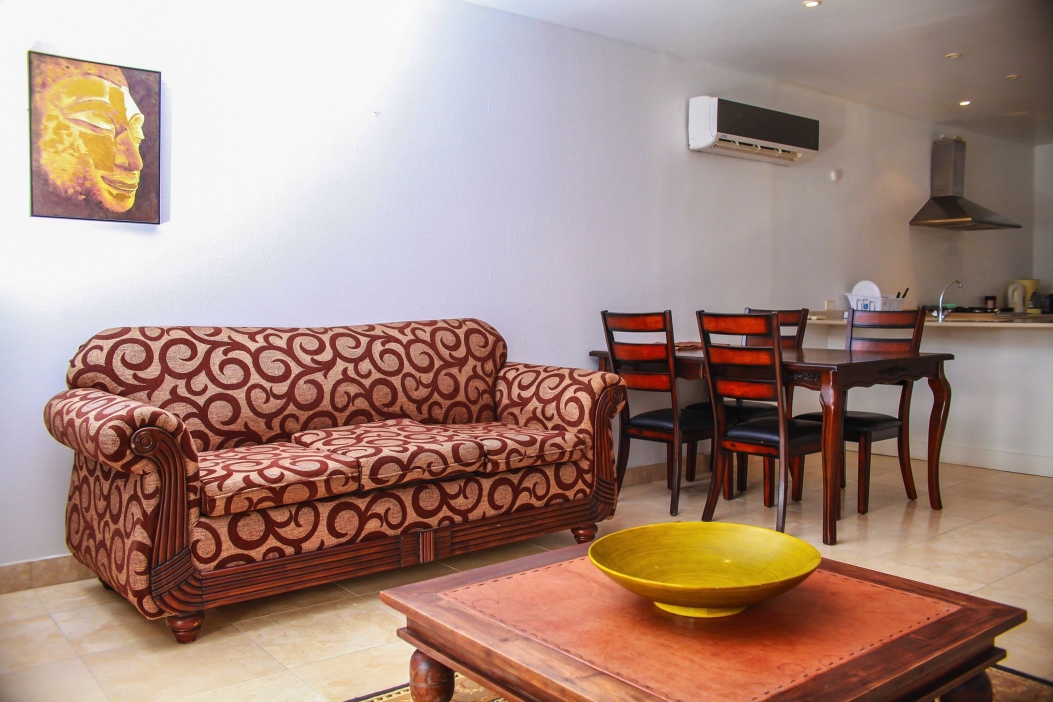 A living room with a patterned sofa, a wooden coffee table with a yellow bowl, and a dining area with chairs and a table.
