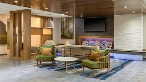 A modern lobby with a wooden ceiling, a flat-screen TV mounted on a textured wall, and a seating area with green upholstered chairs and a round table.
