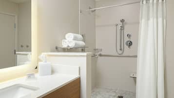 Un baño moderno con un lavamanos blanco, tocador de madera, un espejo, ducha con puerta de cristal y un perchero con toallas dobladas.
