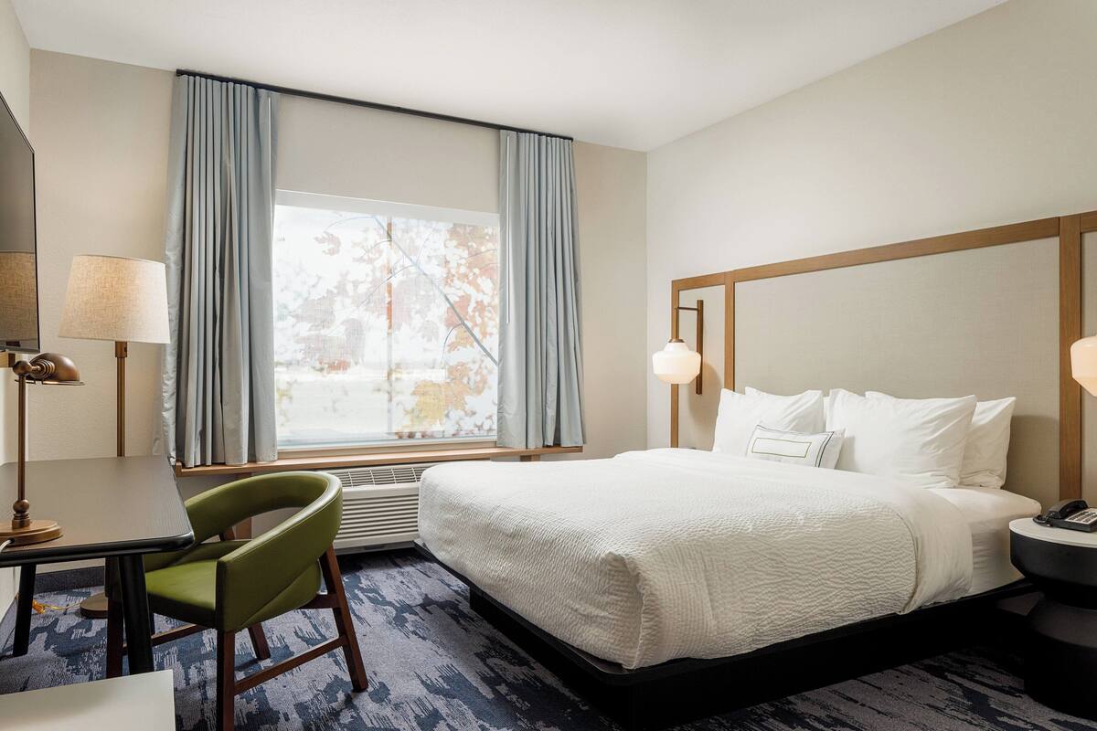 A hotel room with a bed, a desk with a chair, a lamp, and a window with curtains.
