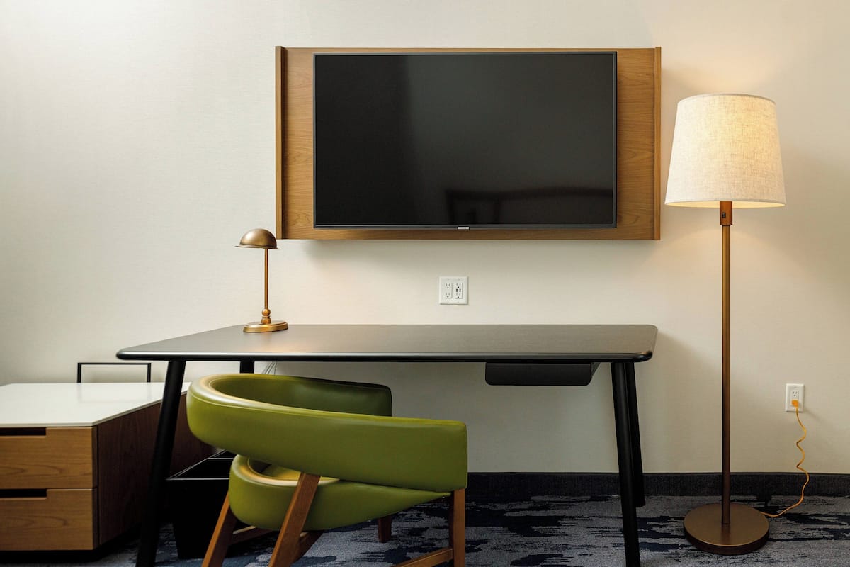 A hotel room with a flat-screen TV mounted on the wall, a desk with a lamp, a green chair, and a floor lamp.