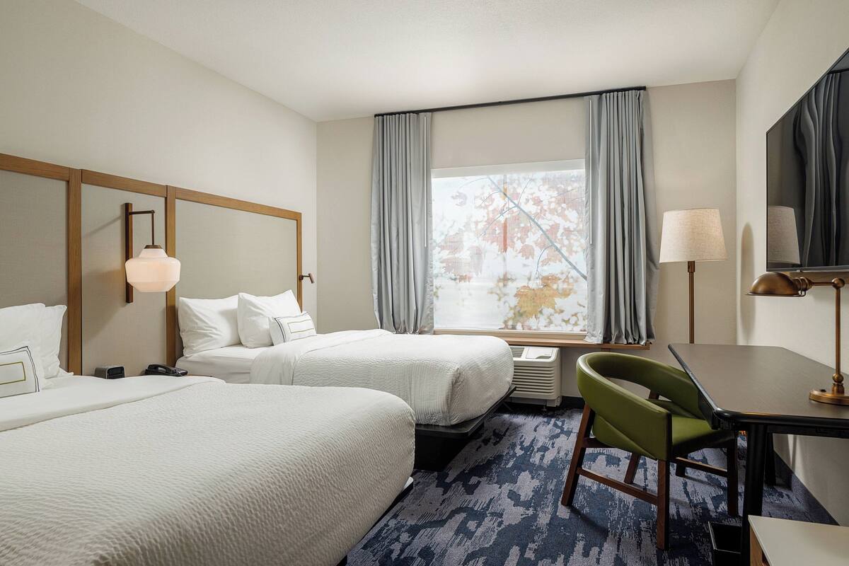 A hotel room with two beds, a desk, a chair, a TV, and a window with curtains.