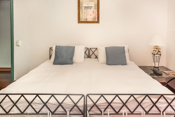 Een netjes opgemaakt bed met een witte deken en twee kussens, een klein nachtkastje met een lamp, en een aan de muur bevestigd fotolijstje.