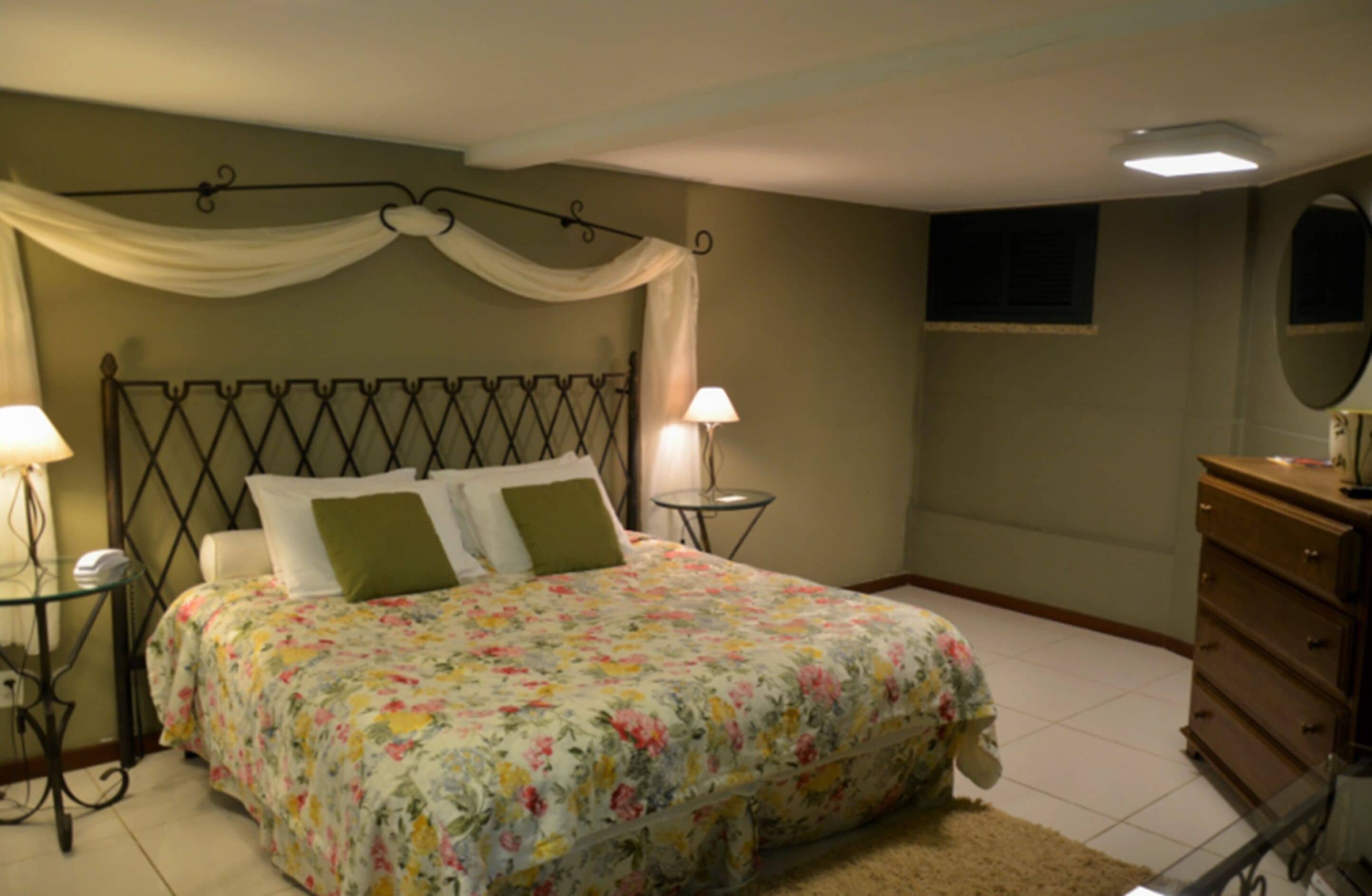Quarto com cama, duas mesinhas de cabeceira, cômoda, espelho e janela com cortinas.