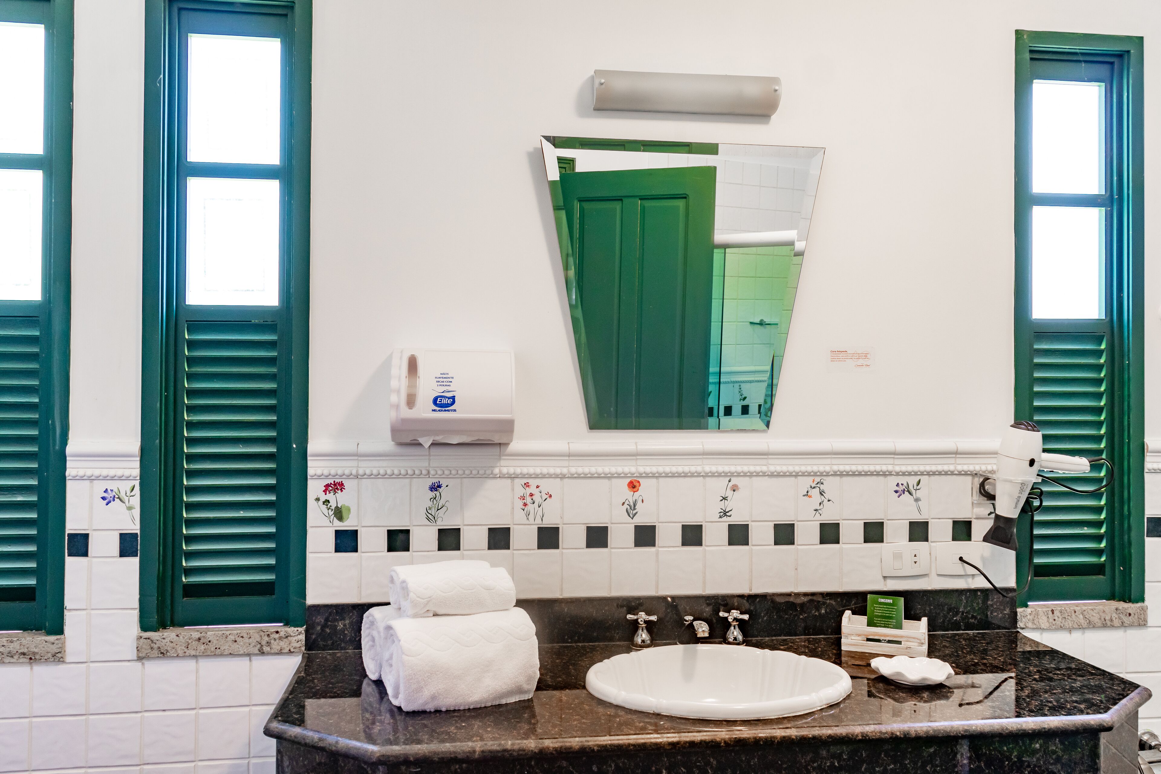 A bathroom with a sink, two towel dispensers, a mirror, and green shutters on the windows.