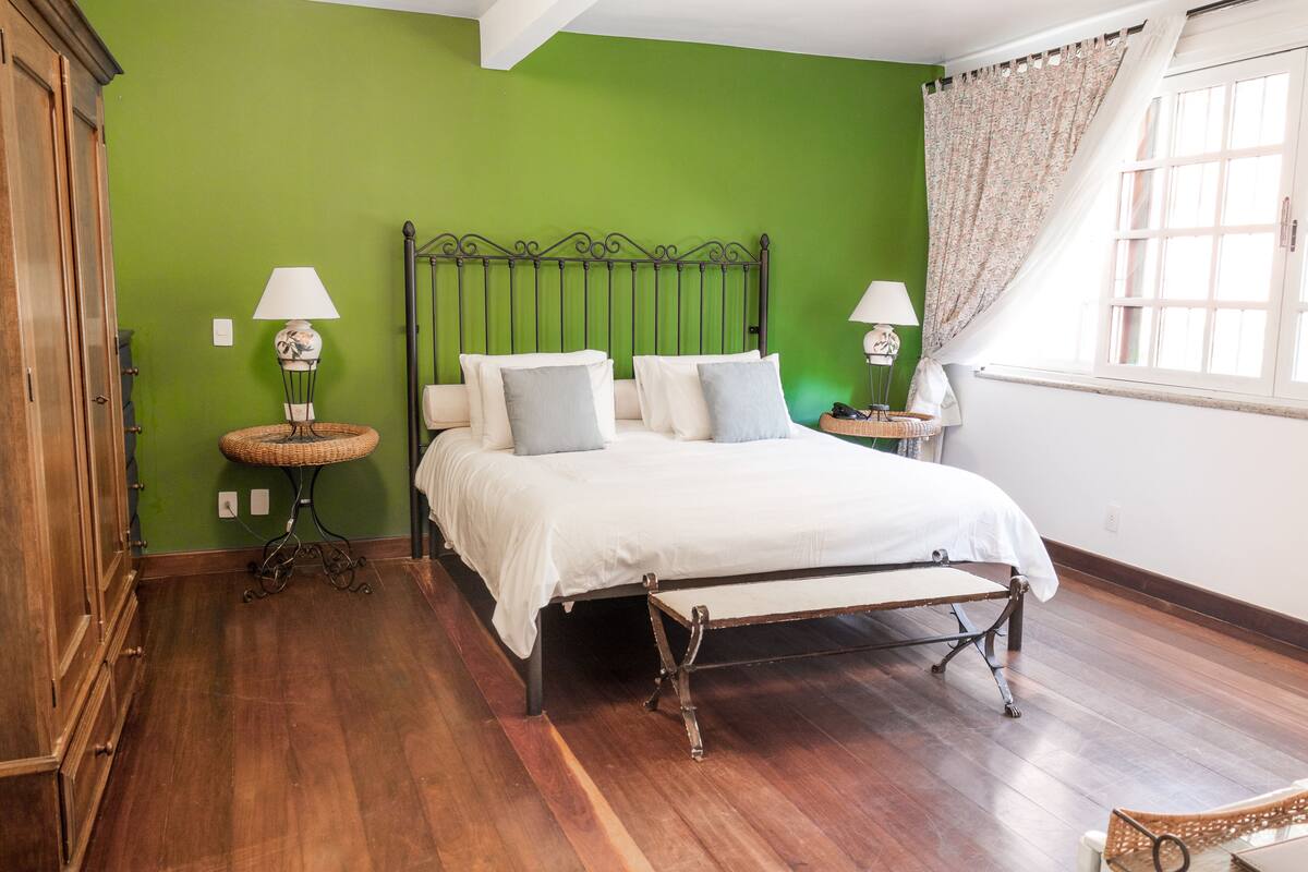 Un dormitorio con pared verde, suelo de madera, estructura de cama metálica, ropa de cama blanca, dos mesitas de noche con lámparas y una ventana con cortinas.