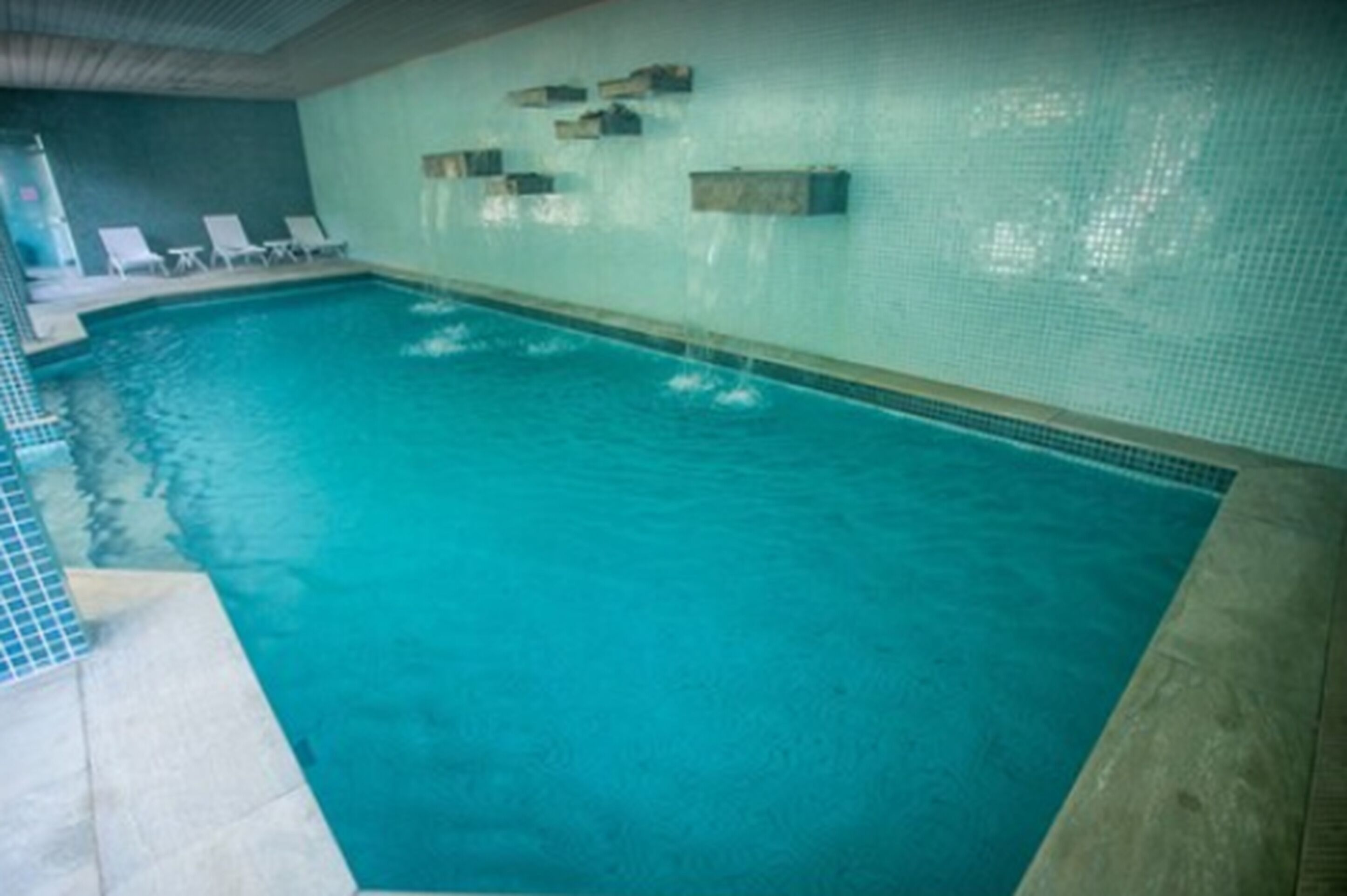 Una piscina interior con agua azul cristalina, paredes con azulejos y algunas sillas de descanso a un lado.