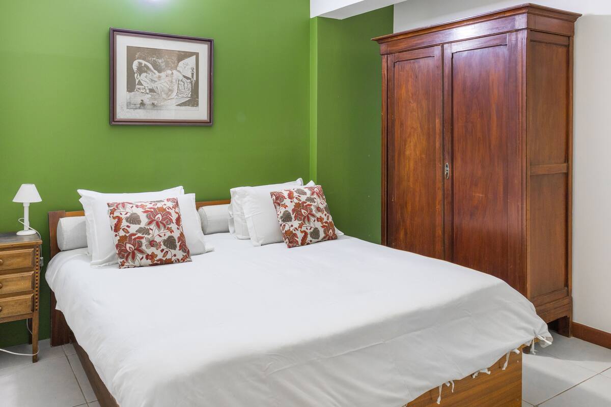 Un dormitorio con cama, almohadas, mesita de noche, lámpara y armario.