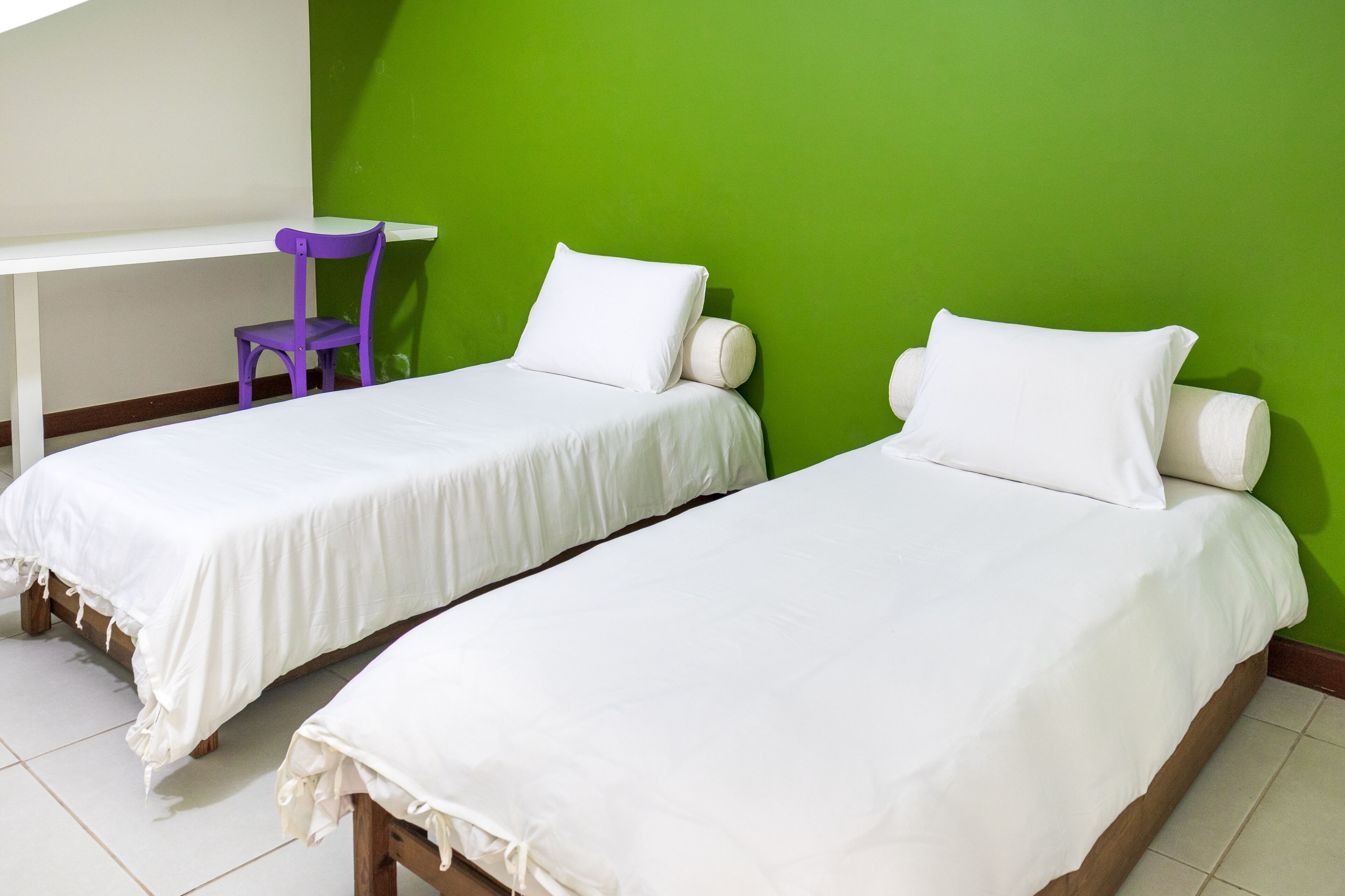 Dos camas individuales con ropa de cama blanca, una silla morada y un escritorio blanco en una habitación con paredes verdes.