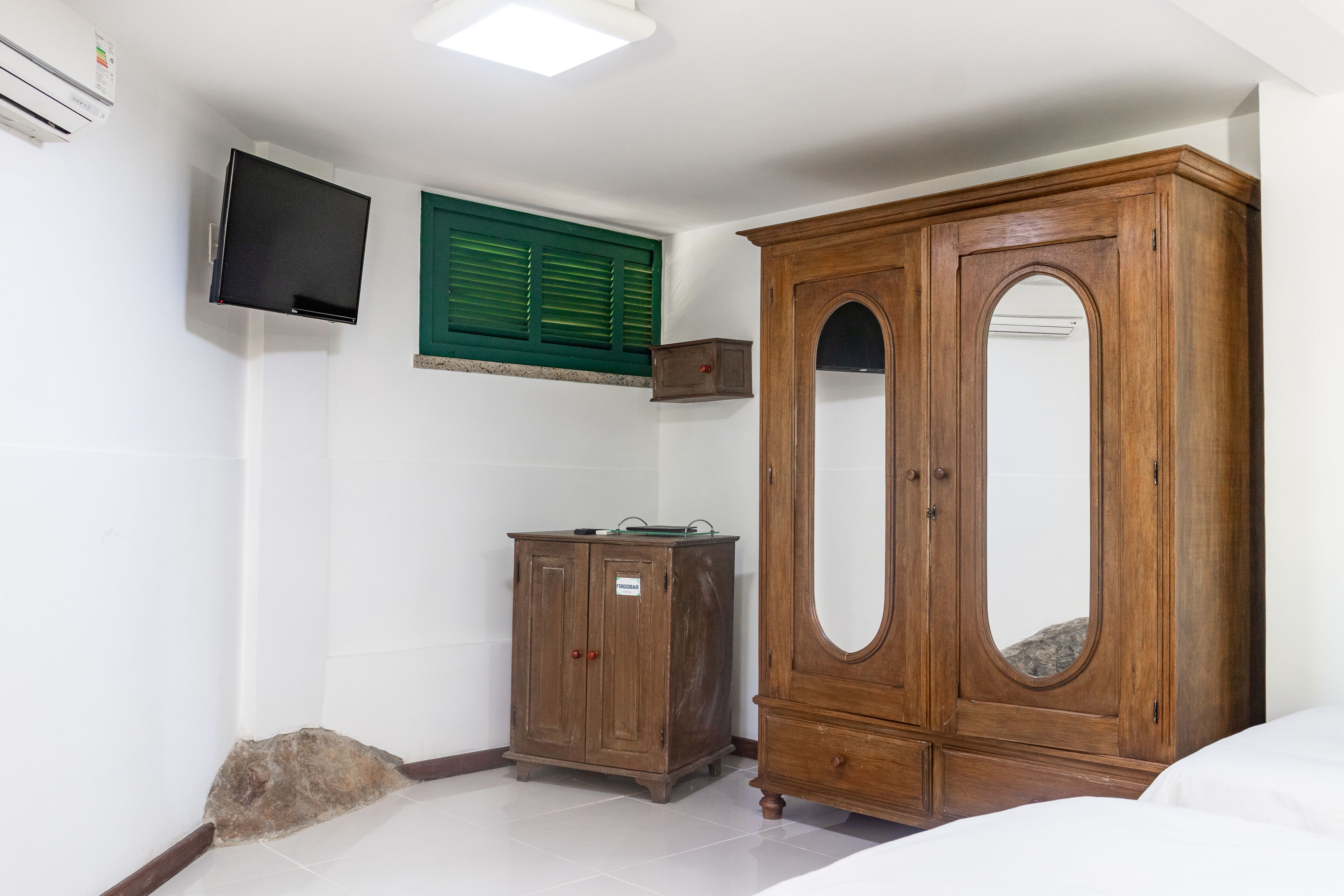 A bedroom with a wooden wardrobe, a television mounted on the wall, and a green window shutter.