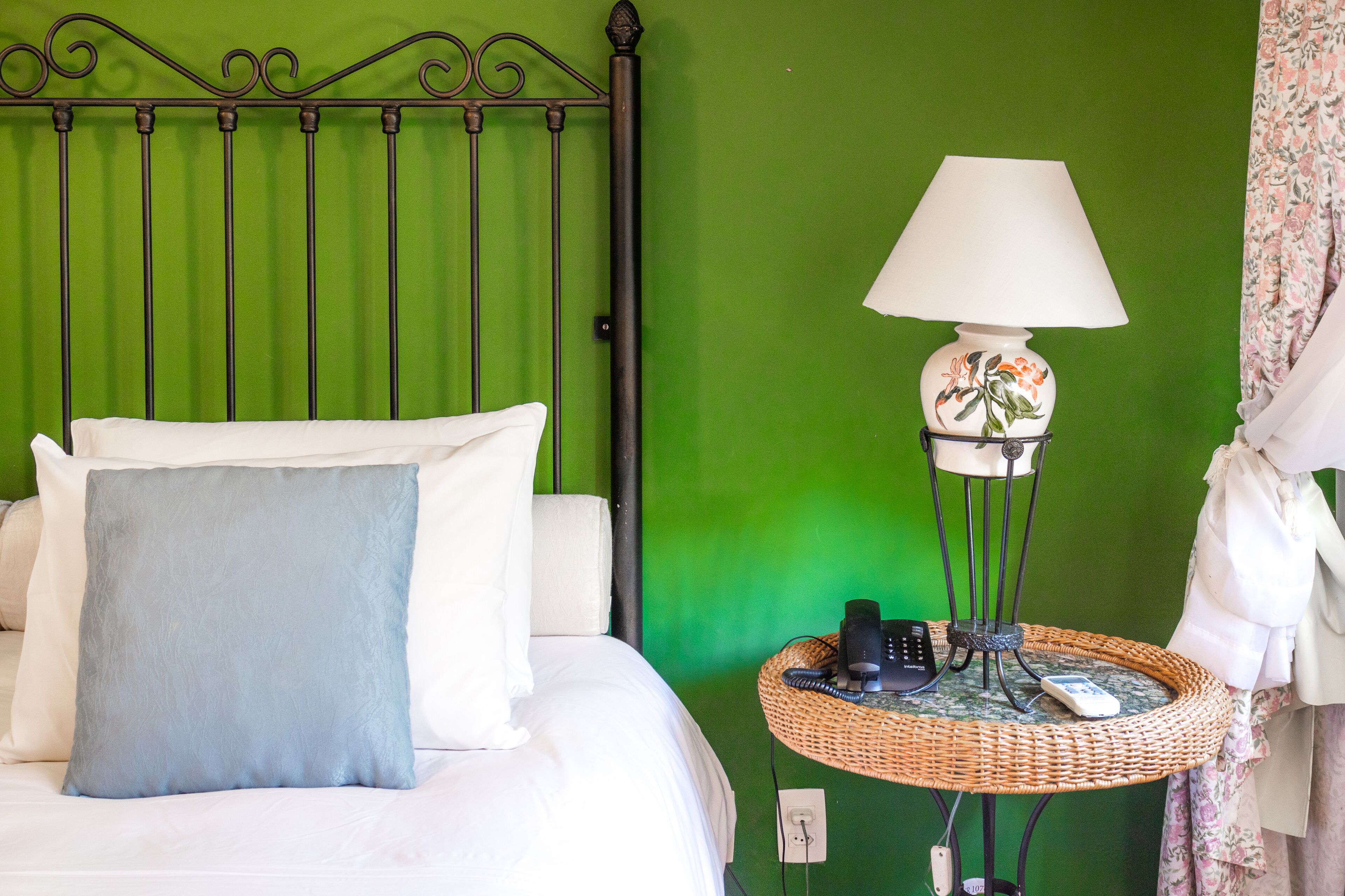 A bed with white and blue pillows, a green wall, a wicker side table with a lamp, and a telephone.