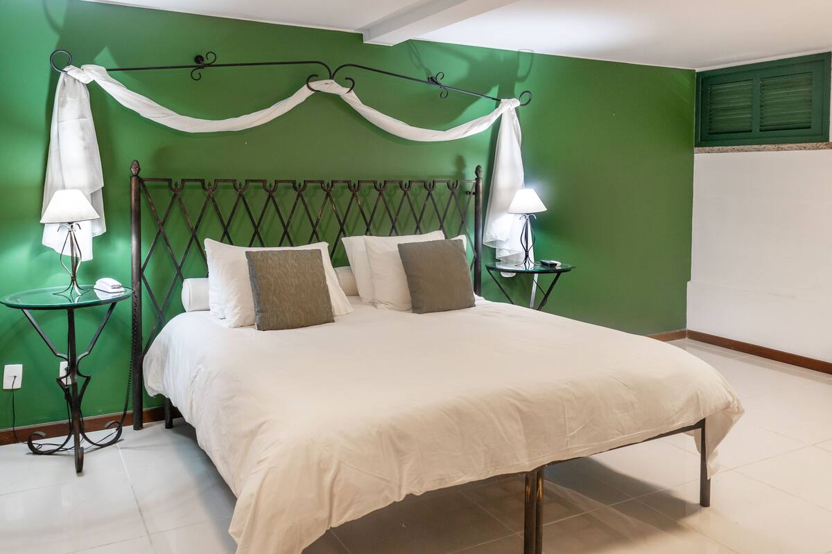Una cama bien tendida con sábanas blancas, dos lámparas de noche, una pared verde y un cabecero de metal.