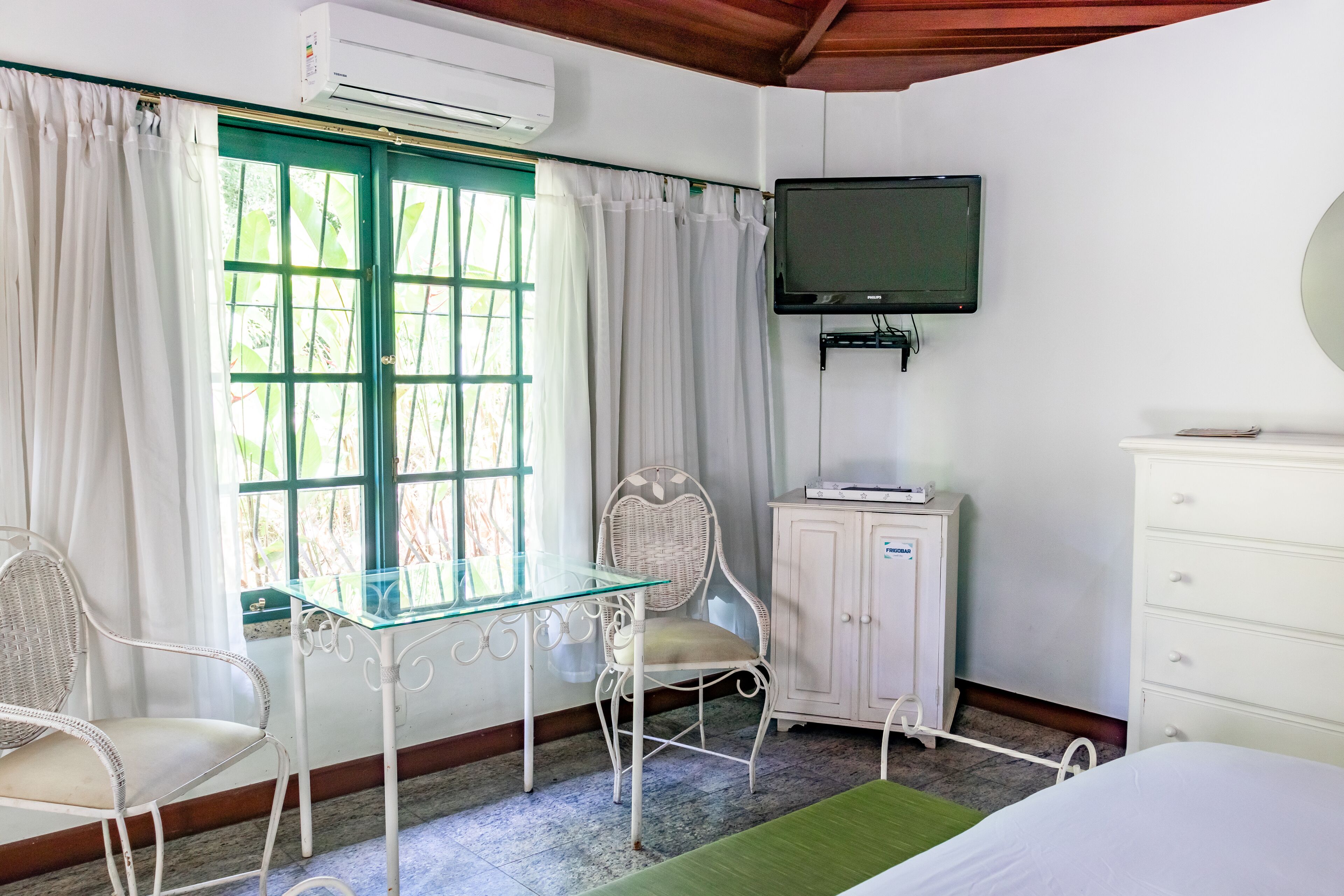 Una habitación con cama, una mesa de vidrio, una silla de mimbre, un armario y un televisor montado en la pared.