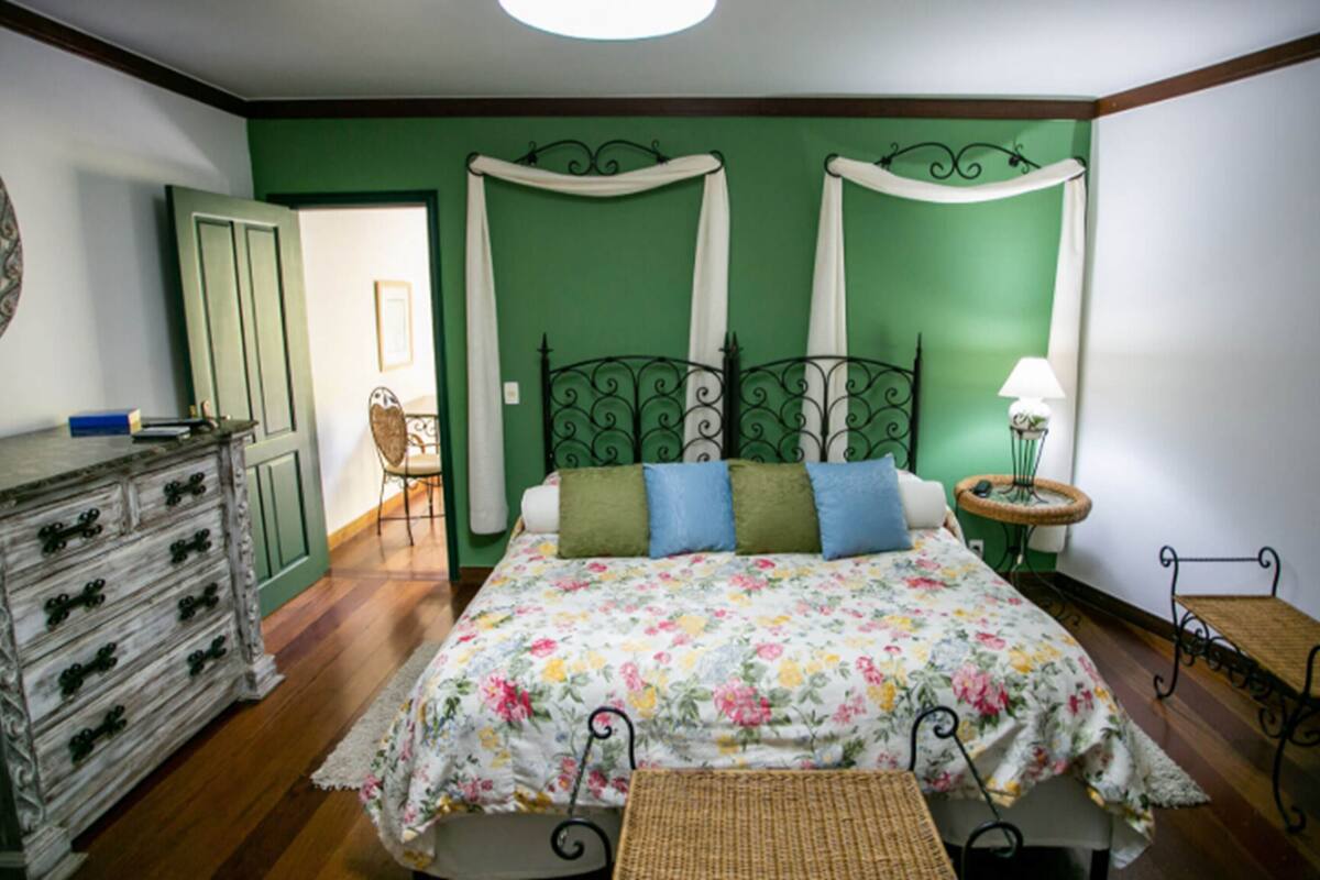Un dormitorio con edredón estampado con flores, cómoda con cajones, una silla, mesita de noche con lámpara y una pequeña mesa con una cesta.