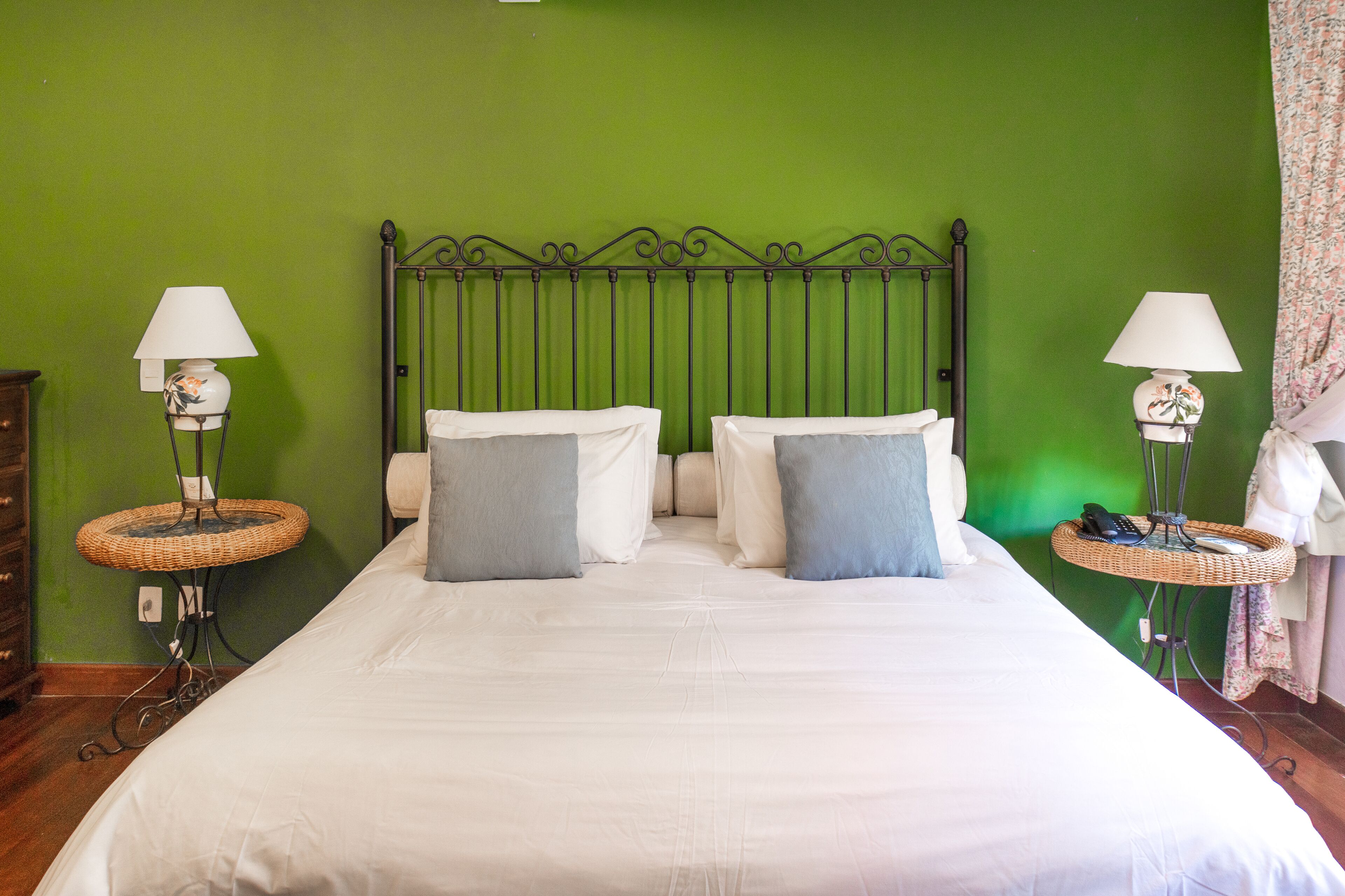 Uma cama de casal com lençóis brancos e duas almofadas, com duas mesinhas de vime e abajures laterais, num quarto com paredes verdes.