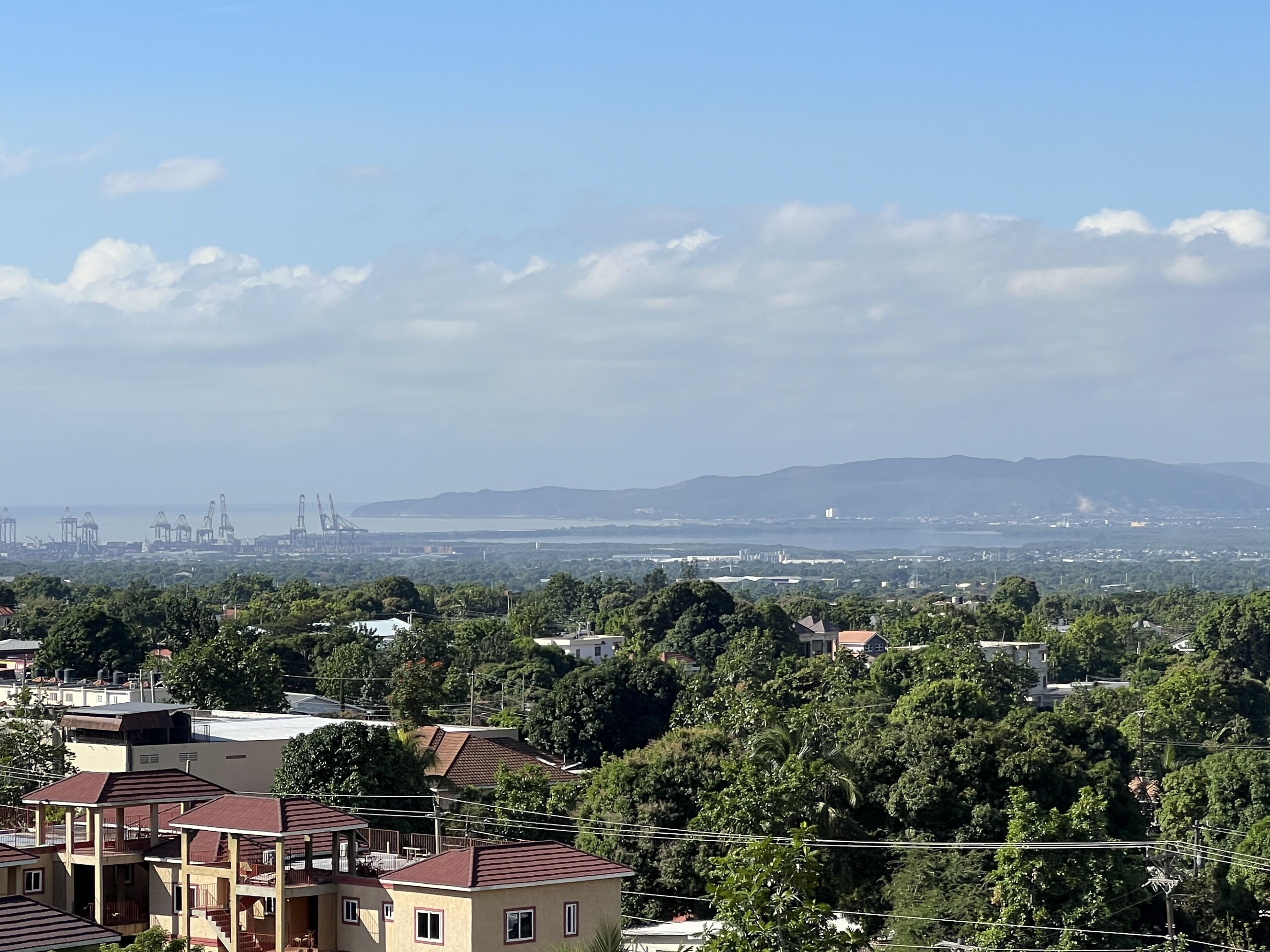 A residential area with multi-story buildings, dense greenery, and a distant view of a port with cranes.