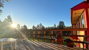 Eine Holzterrasse mit Tisch und Stühlen, ein rotes Gebäude und im Hintergrund Bäume sowie ein Berg.