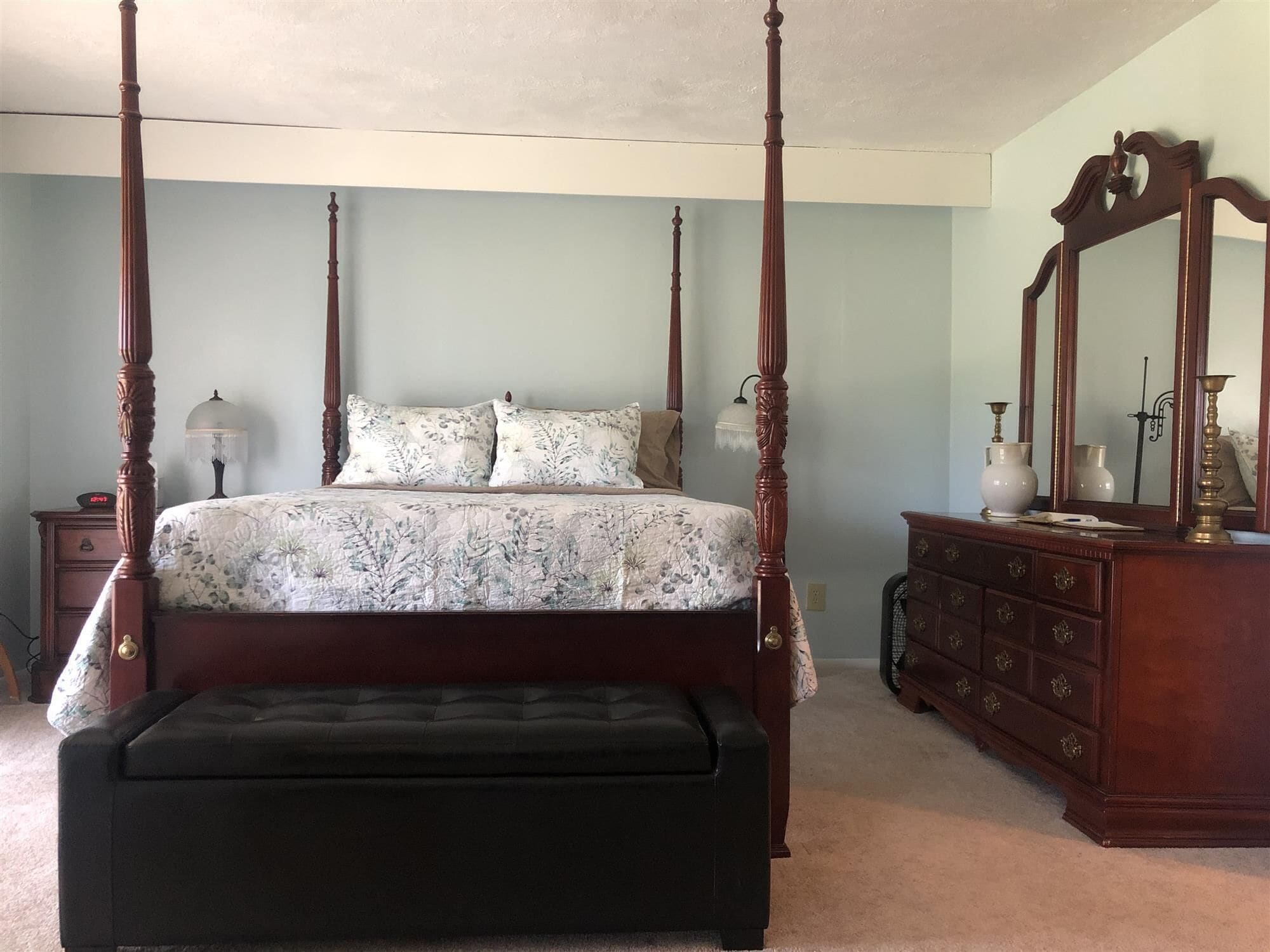 A four-poster bed with a floral bedspread, a wooden dresser with a mirror, and a small bedside table with a lamp.