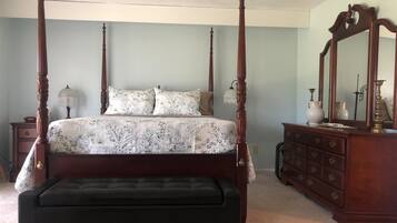 A four-poster bed with a floral bedspread, a wooden dresser with a mirror, and a small bedside table with a lamp.