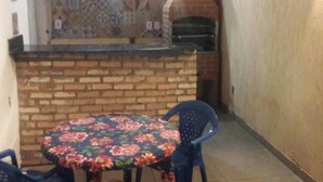 A small patio area with a brick oven, a round table with floral tablecloth, and blue plastic chairs. - Casa com Lazer Completo (Cidade Ocidental)