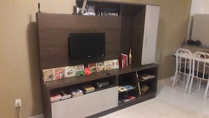 A living room with a TV mounted on a wooden cabinet, a small shelf with books and decorative items, and a white chair in the corner. - Casa com Lazer Completo (Cidade Ocidental)