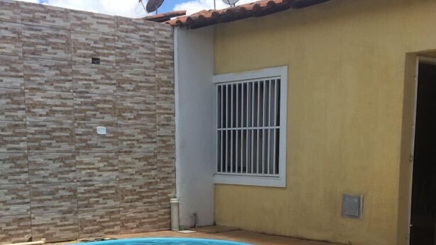 A swimming pool with a tiled deck, a brick wall, and a building with a tiled roof and a barred window. - Casa com Lazer Completo (Cidade Ocidental)