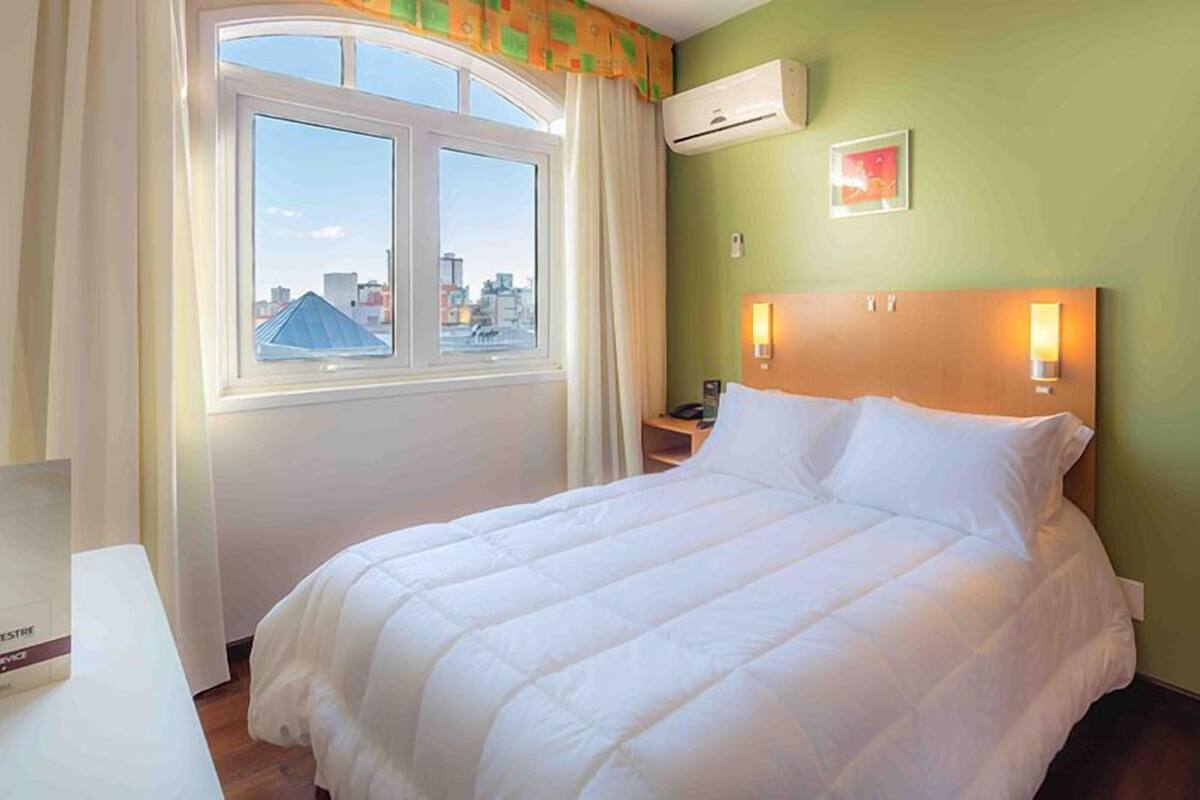 Une chambre d’hôtel comprenant un lit, une table de chevet, un appareil de climatisation et une fenêtre donnant sur la ville.