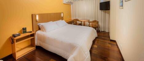 Habitación de hotel con una cama grande, una mesita de noche con teléfono, un pequeño escritorio con silla, un televisor montado en la pared y una ventana con cortinas.