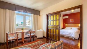 A hotel room with a bed, a dining area with a table and chairs, a view of the city through the window, and a red accent wall.