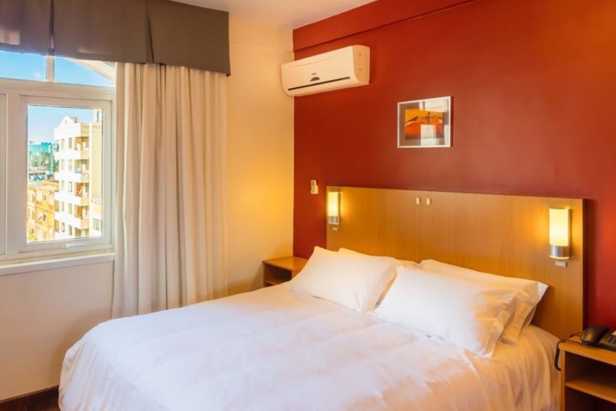 Une chambre d’hôtel avec un grand lit, un appareil de climatisation, une table de chevet avec un téléphone et une fenêtre avec des rideaux.