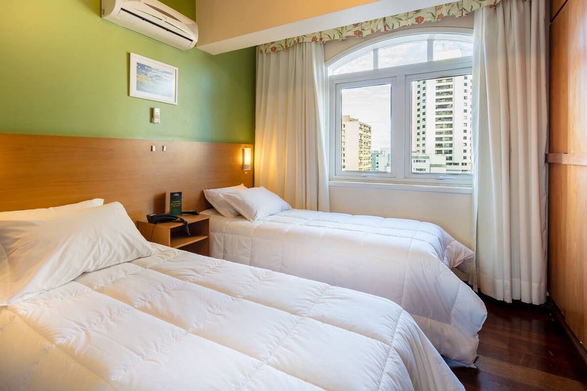 Une chambre d’hôtel avec deux lits, une table de chevet, un climatiseur fixé au mur, un tableau encadré et une fenêtre avec des rideaux.